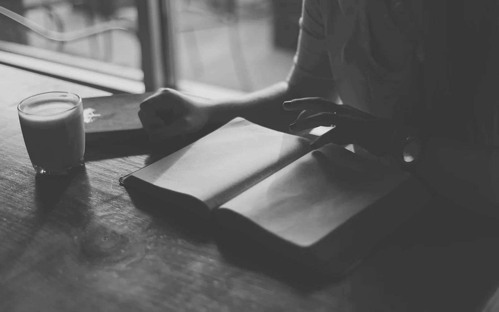 Una persona lee un libro junto a una taza de café y un cuaderno.