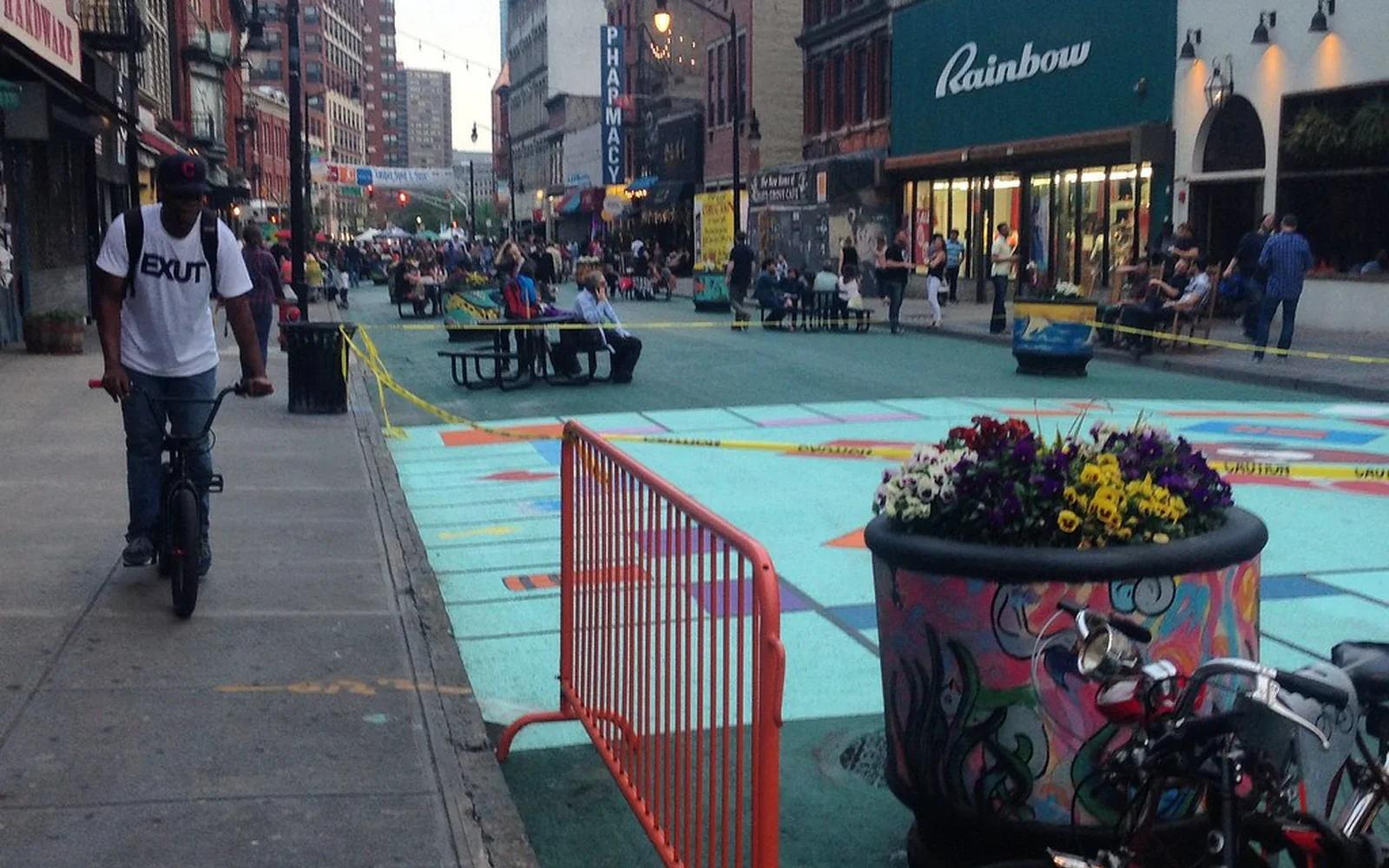 Zona peatonal de una ciudad con bancos, árboles y personas caminando.
